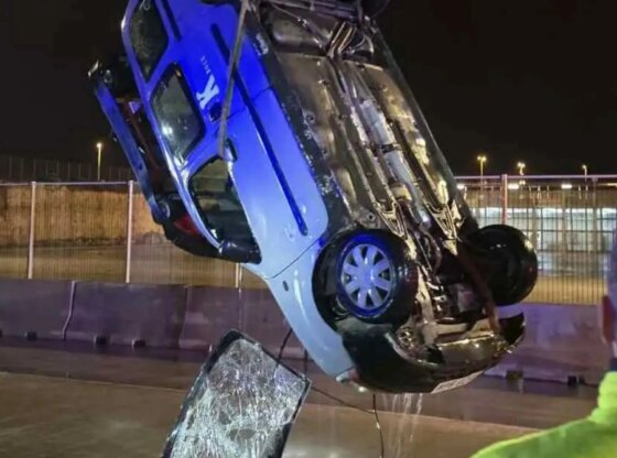 An image collage containing 1 images, Image 1 shows A blue and white car being lifted upside down by a crane from the water at night, with a person in a safety vest watching