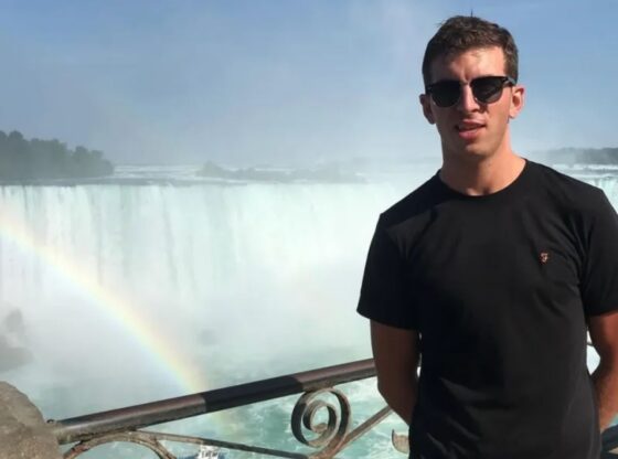 An image collage containing 1 images, Image 1 shows Man in sunglasses and black shirt standing in front of Niagara Falls with a rainbow and a boat below