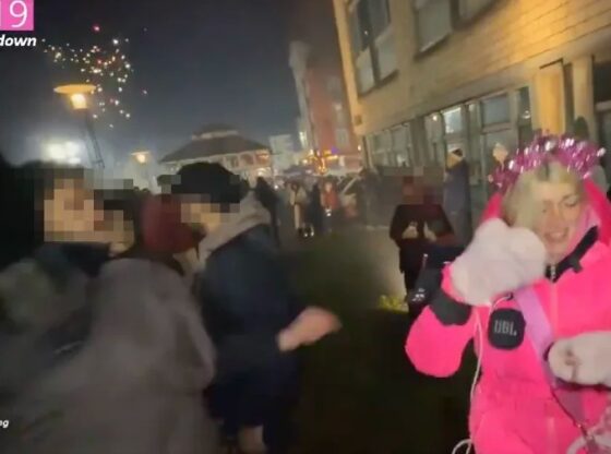 A woman in a pink jacket and festive crown being attacked by a firework during a New Year's Eve countdown.