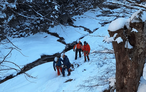 Four people in winter gear and helmets navigating a snow-covered landscape next to a frozen stream.